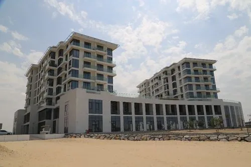 A modern, multi-story building with large windows and balconies, featuring two connected structures. The building is set against a partly cloudy sky, and a sand-covered area is visible in the foreground, bordered by a low stone wall.